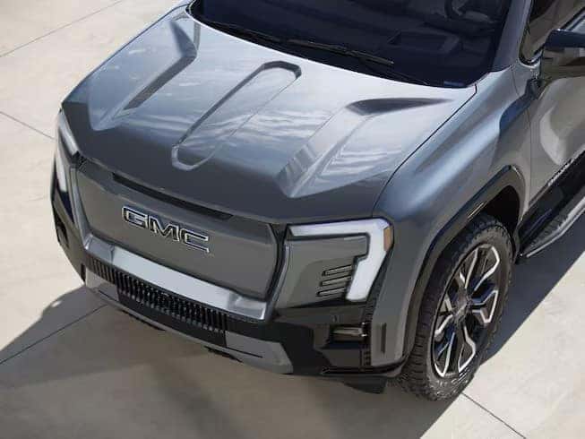 Close-up view of a sleek, gray GMC truck. The vehicle features a modern design with distinctive LED headlights, a bold grille displaying the GMC logo, and rugged tires. The truck is parked on a light-colored concrete surface.