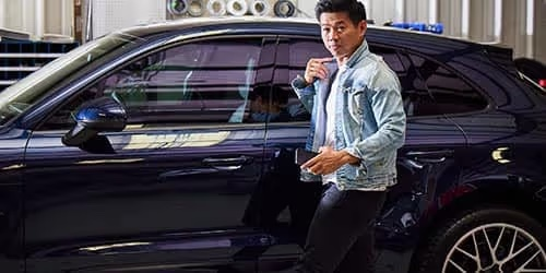 A man wearing a light denim jacket stands next to a dark-colored car in a garage, looking toward the camera. The car’s door is closed and various equipment is visible in the background.