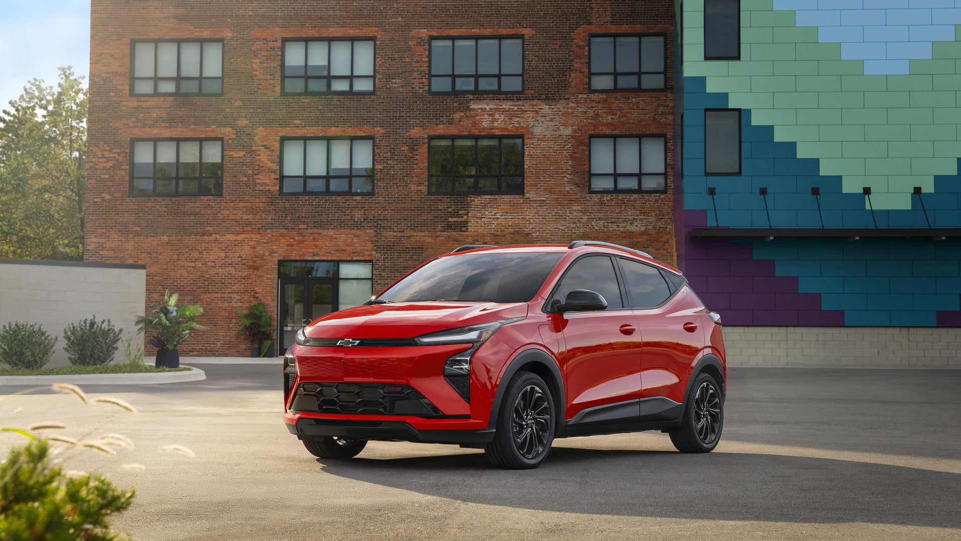 A red SUV with black and chrome accents is driving on a city street. Its a four-door vehicle with large alloy wheels, a sleek design, and tinted windows. The background features blurred buildings and trees.
