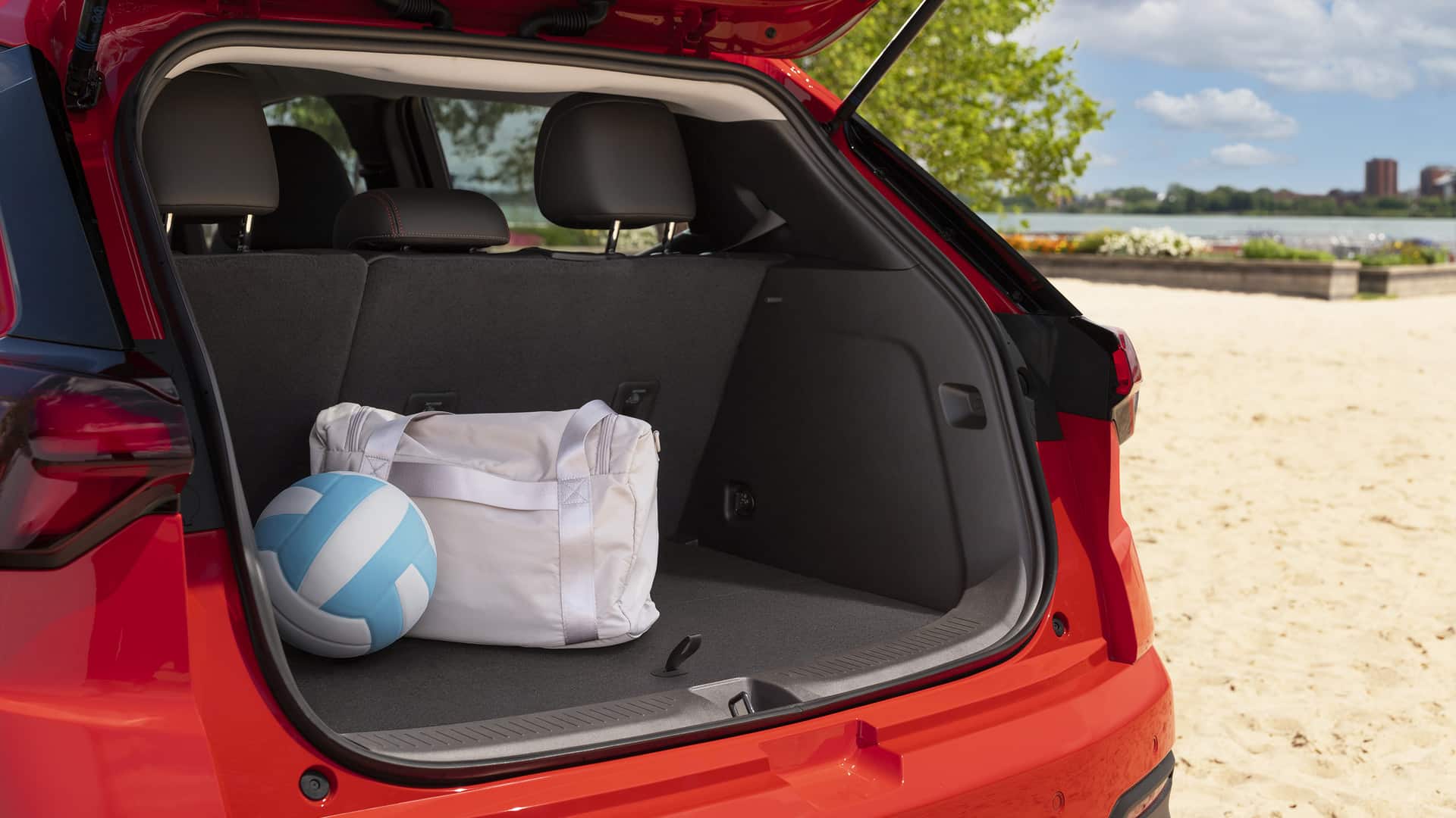 Open trunk of a red car at the beach with a sports bag and volleyball inside.