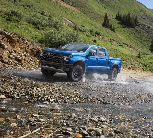 A blue pickup truck driving through a shallow stream with a lush green hillside in the background.