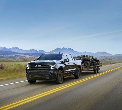 A black pickup truck towing a trailer on a scenic highway with mountains in the background under a clear sky.