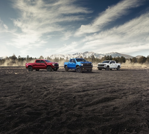 Three pickup trucks in red, blue, and white on a rugged landscape with mountains and cloudy sky.