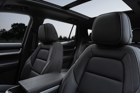 View from the backseat of a car showing two people in the front seats. The driver is holding the steering wheel. The sunroof is open, revealing a blue sky and tall buildings outside. The dashboard and rearview mirror are visible.