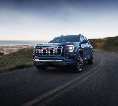 A sleek SUV driving on a winding road with coastal scenery in the background under a clear sky.