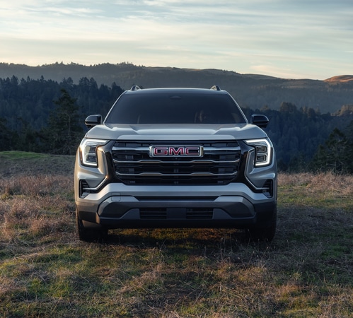 Front view of a GMC truck parked on grassy terrain with forested hills in the background at dusk.