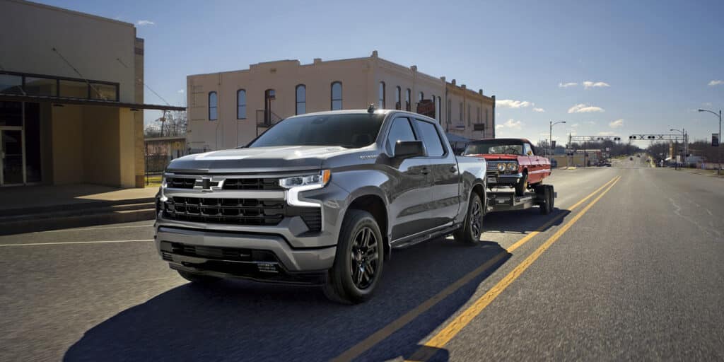 Silver pickup truck towing a classic red car on a city street under a clear blue sky.