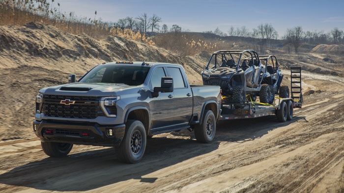 A blue pickup truck towing a trailer with two off-road vehicles on a rugged dirt path.