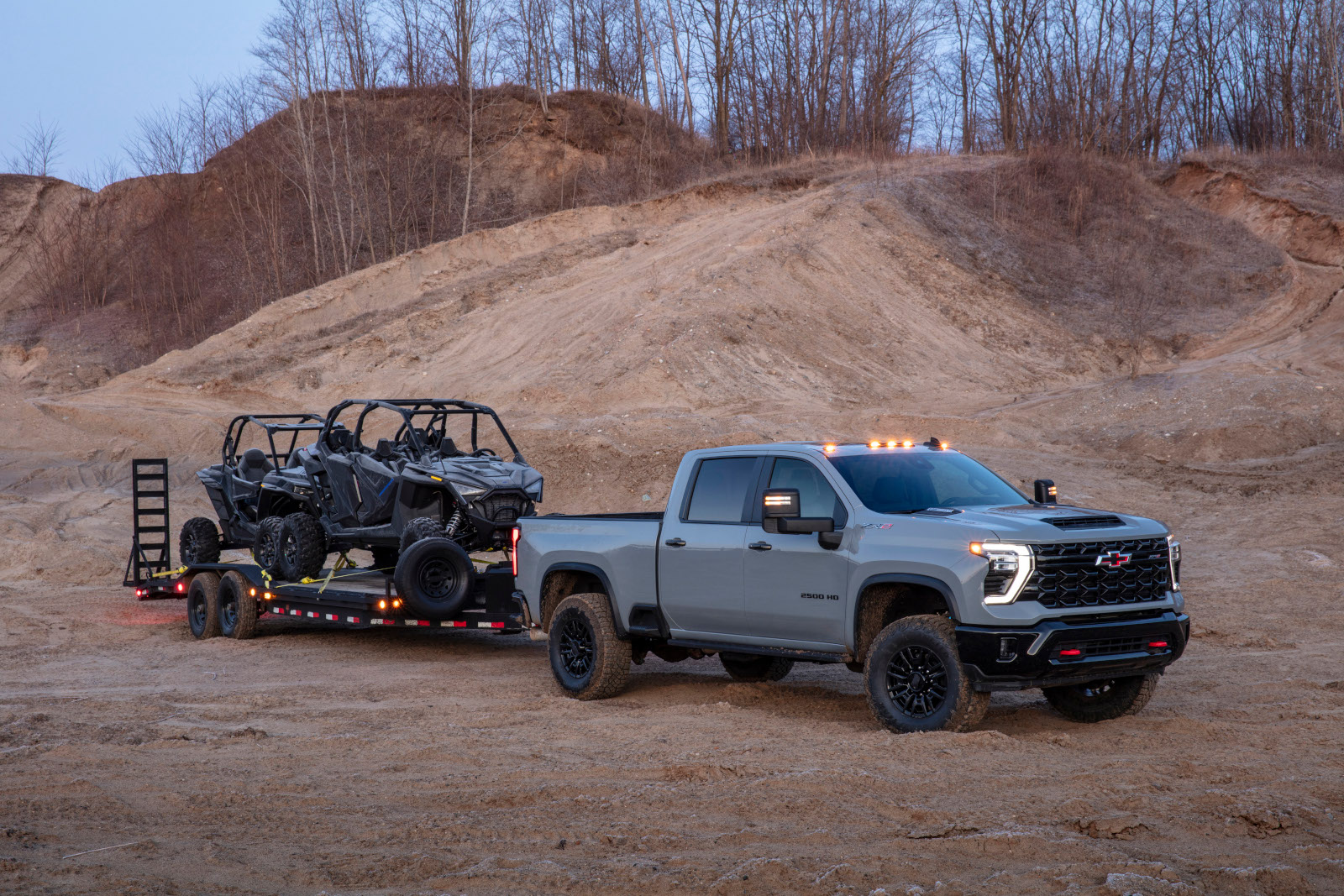 Truck towing off-road vehicles on a trailer in a sandy landscape with bare trees.