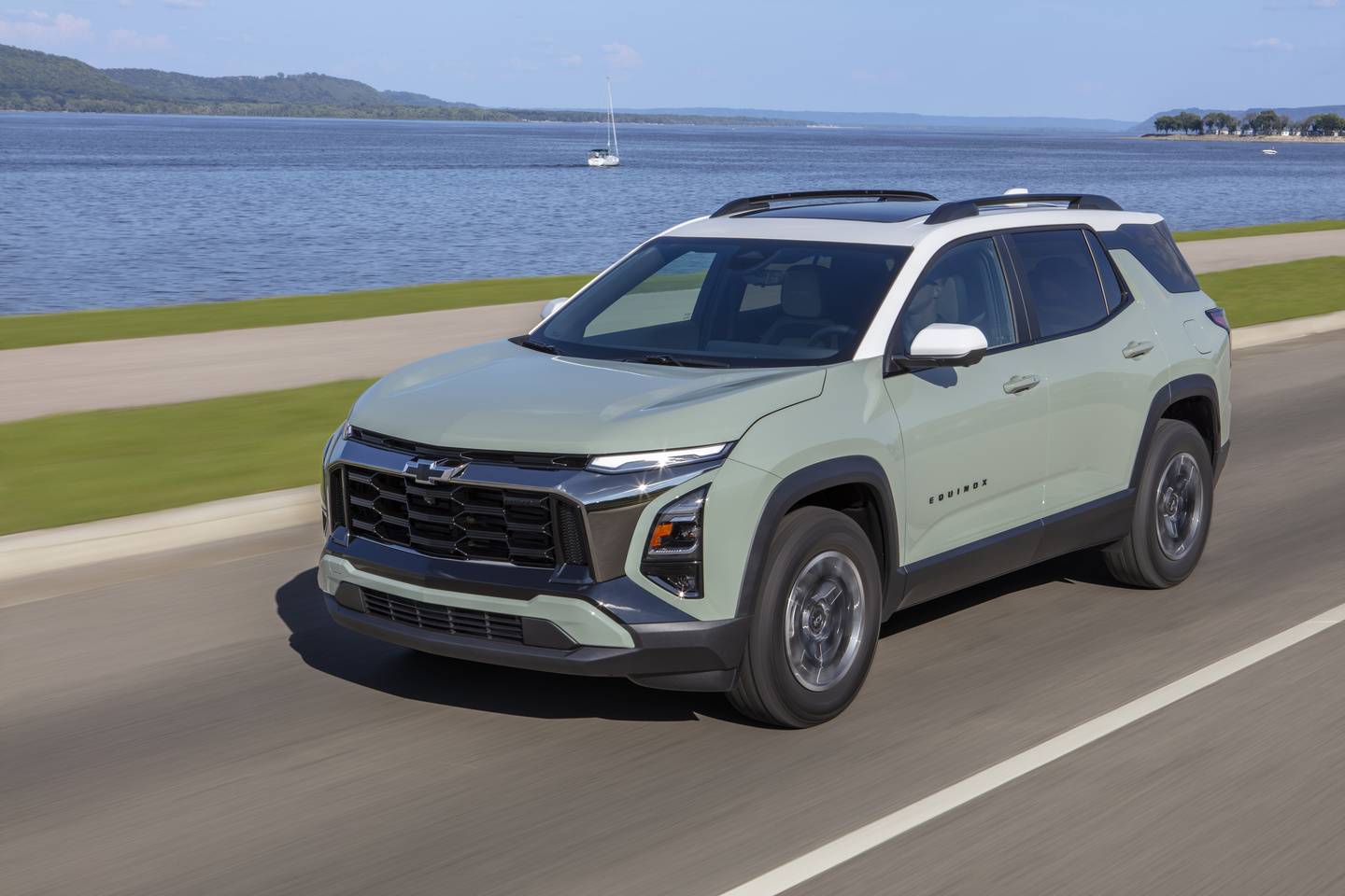 A light green SUV driving on a coastal road with a scenic water view in the background.