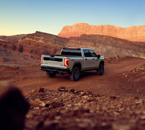 A pickup truck drives on a dirt road through a rugged desert landscape at sunset.