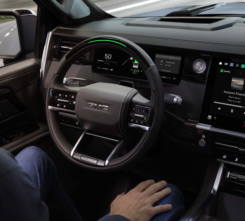 Driver in a GMC vehicle with advanced tech displays on the dashboard, showing speed and music info.