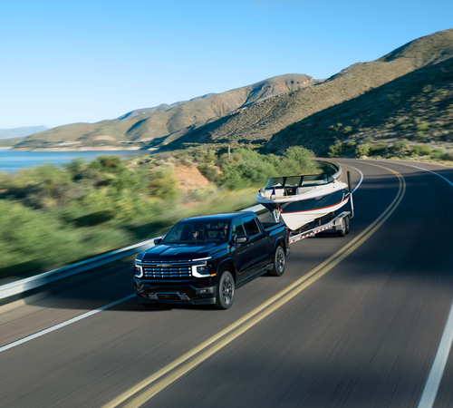 Truck towing a boat along a scenic lakeside highway, with mountains in the background under a clear blue sky.
