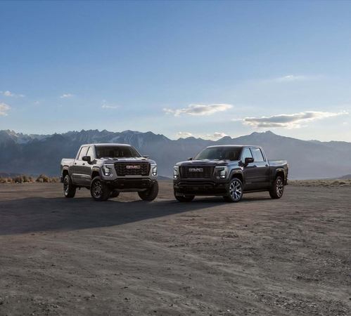 Two GMC trucks parked on a dirt road with a mountainous landscape and blue sky backdrop.